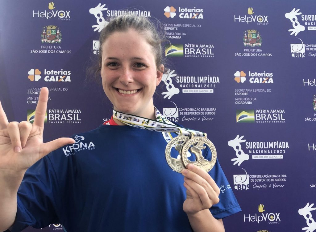 Mariana de Andrade de Mello aparece na foto, posicionada do centro à esquerda, mostrando suas medalhas de prata conquistadas em 2021. Ela é uma mulher branca, de cabelo castanho-claro, que está preso em um coque, e olhos escuros. Ela está sorrindo e fazendo o sinal "Amo você" em língua de sinais. Ao fundo, está um backdrop/painel azul com os patrocinadores da competição nacional de surdolimpíada.